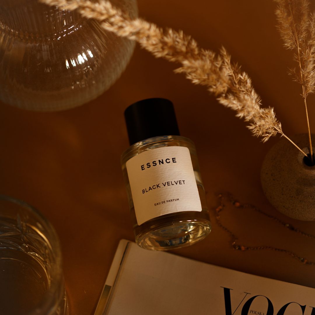 En flaska ESSNCE Black Velvet Eau de Parfum med en svart kork är placerad på en brun yta bredvid en bukett med torkat pampasgräs, en glasbehållare och en VOGUE-tidning. Kompositionen utstrålar en varm, sofistikerad atmosfär.