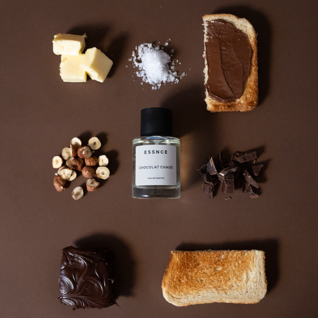 En texturerad brun yta med ett urval av chokladbitar, nötter och rostade brödskivor. En parfymflaska ESSNCE Chocolat Chaud Eau de Parfum ligger i mitten.