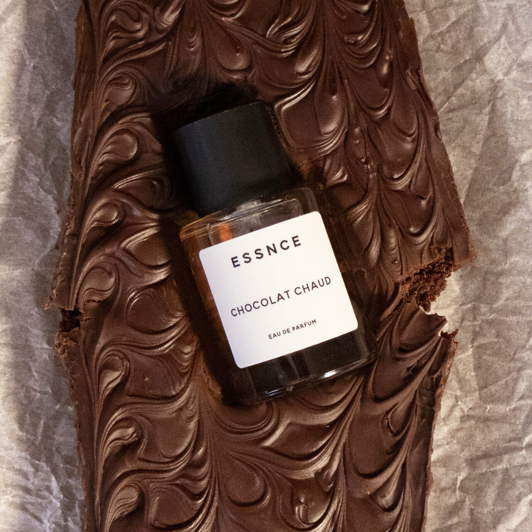 En vacker chokladkaka med en parfymflaska märkt ESSNCE Chocolat Chaud Eau de Parfum i mitten, som framhäver dess lyxiga utseende och inbjudande textur.