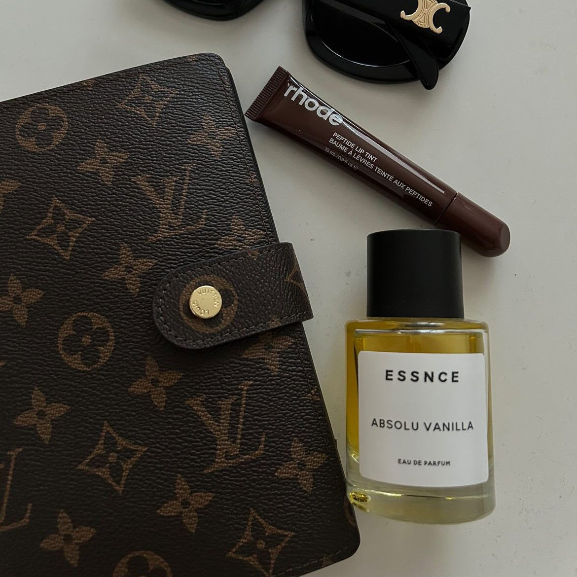 En flatlay-bild visar en brun Louis Vuitton-plånbok, ett par svarta solglasögon med gulddetaljer, en bruntonad tub av rhode lip tint och en flaska ESSNCE Absolu Vanilla Eau de Parfum.