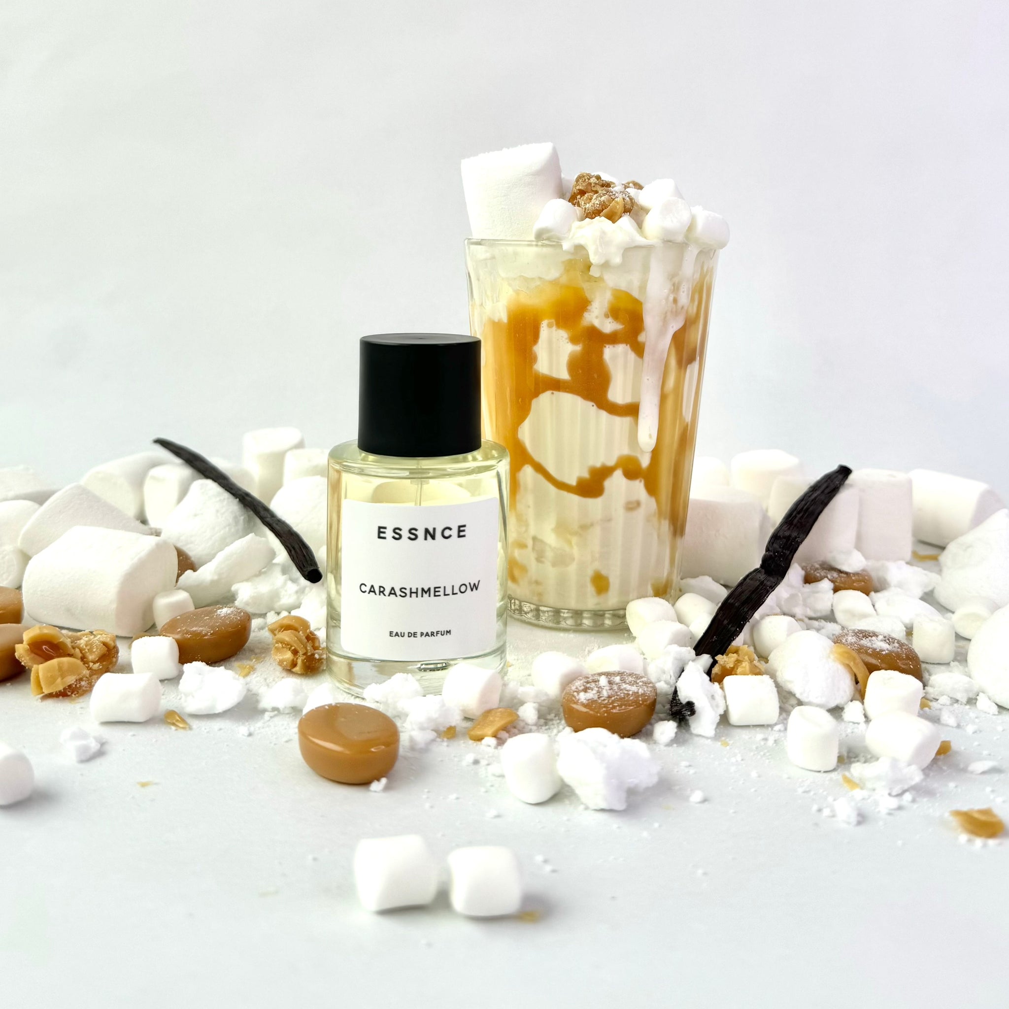 En flaska ESSNCE Carashmellow Eau de Parfum visas med en svart kork och en vit etikett. Parfymen är omringad av massor av marshmallows, kola, och ett glas med milkshake med karamell i.