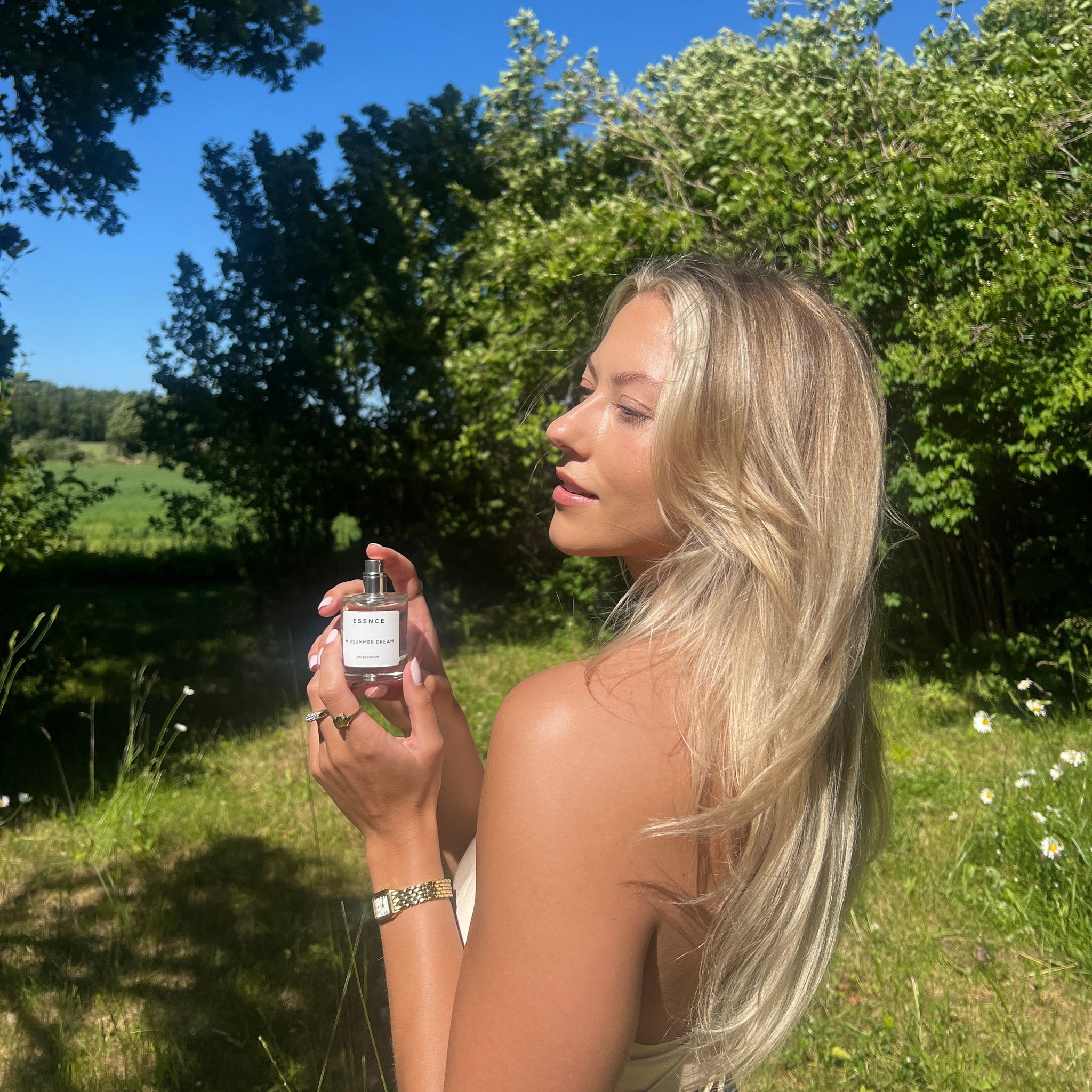 En flaska ESSNCE Midsummer dream Eau de Parfum som hålls up av någon som står på en grön trädgård på sommaren.