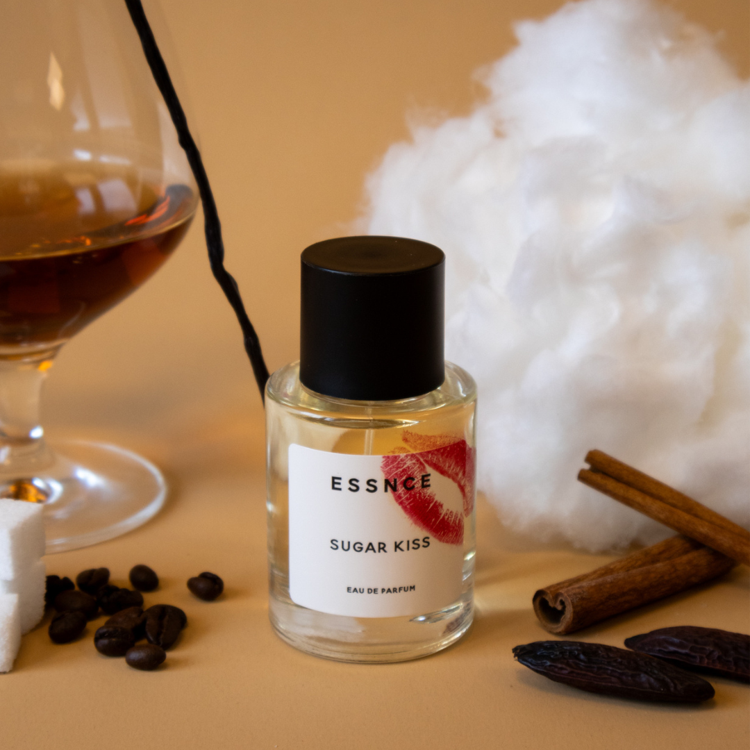 En flaska parfym med etiketten ESSNCE Sugar Kiss Eau de Parfum står placerad intill ett glas rom, sockervadd, kaffebönor, sockerbitar, vaniljstänger och kanelstänger. Parfymflaskan har ett rött läppsticksmärke på sig, vilket ger en känsla av lyx och värme.