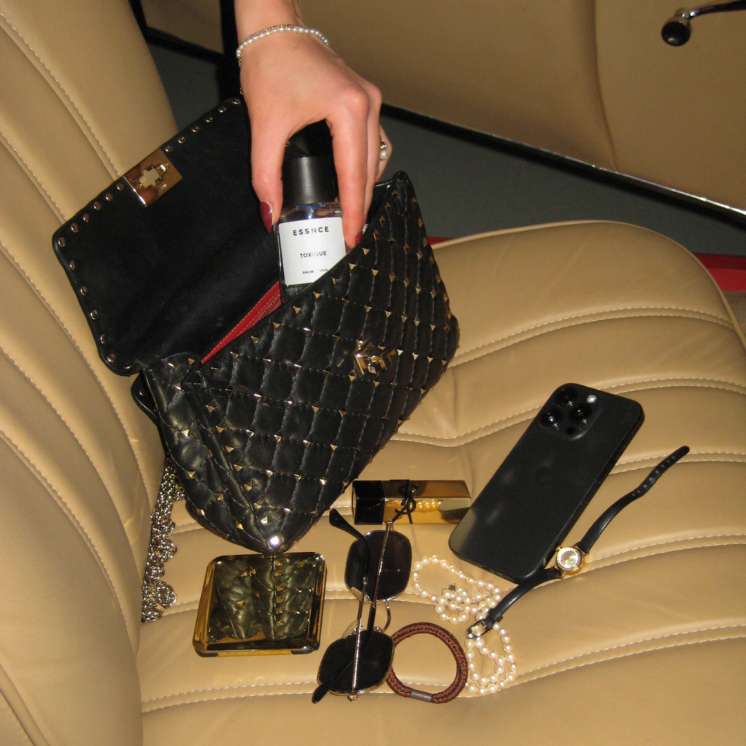 A hand is seen taking a bottle of Toxique perfume out of a black quilted bag on a beige leather seat in a car. Around the bag, various items are scattered, including a smartphone, a wristwatch, sunglasses, a compact mirror, lipstick, and a pearl necklace.