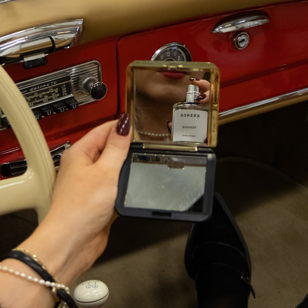 A person is holding a small mirror with a Toxique perfume bottle in front of their face while sitting in an old car. The car's dashboard and steering wheel are partially visible.